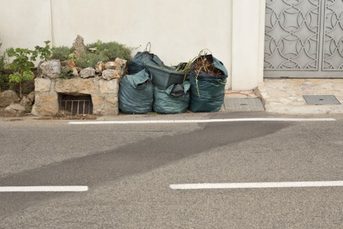 A row of four large black garbage bags filled with waste material, placed on the edge of a pavement in front of a white wall with a small garden bed containing green plants and rocks on the left, and a decorative metal gate on the right. The bags are slightly bulging and tied at the top, positioned close together along the curbside, with an asphalt road running parallel in the foreground. The scene suggests a rubbish collection or disposal area, typical of commercial waste removal services operating in Tottenham, London, with the bags prepared for collection or disposal by Business Waste Removal Tottenham.