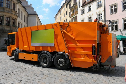 A large, modern orange rubbish collection truck is parked on a cobbled street in a historic area, with multi-storey white and cream-colored buildings visible in the background. The vehicle features a smooth, shiny metallic body with a prominent rectangular green slip on its side, and has multiple wheels and heavy-duty tires designed for urban waste collection. The rear section of the truck is equipped with hydraulic arms and a waste compaction mechanism, indicating it is prepared for efficient rubbish removal. The scene suggests a planned street cleaning or waste collection operation typical of urban waste management services in Tottenham, with clear natural daylight illuminating the environment. The background buildings have ornate facades and large windows, reflecting the architectural character of the area. Business Waste Removal Tottenham would handle such waste collection needs in the local postcode area, ensuring debris is properly managed and removed from commercial or residential settings.