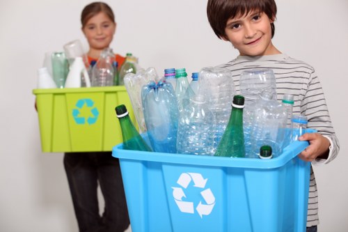 In the foreground, a young boy with dark hair and a striped grey and white shirt is holding a blue plastic recycling bin filled with empty plastic bottles and containers, including clear and green-tinted bottles, some with caps and others without. Behind him, a young girl with light brown hair and a grey top is holding a light green recycling bin with a visible blue recycling symbol, and is also filled with various plastic bottles and containers. Both children are standing against a plain white background, which emphasizes the presence of the bins. The setting appears to be indoors, possibly in a home or community space, designed to highlight waste separation and recycling activities. The image connects to waste management services such as those offered by Business Waste Removal Tottenham, focusing on the collection and proper disposal of recyclable plastic materials commonly managed in commercial or domestic contexts in Tottenham, London, with clear visual cues of plastic waste sorting and collection.