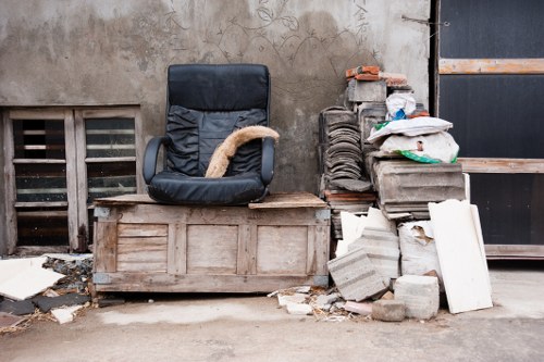 A black office swivel chair with a textured fabric surface and a beige cushioned seat is placed on a weathered wooden chest situated outdoors against a grey concrete wall. To the right of the chair, there is a pile of construction debris consisting of several stacked bricks, concrete blocks, and loose pieces of masonry, some of which are damaged or broken. A white plastic bag and a green item are visible on top of the debris, indicating miscellaneous waste. The debris stack is slightly leaning against a dark wooden panel or shelving unit partially visible on the far right. The ground appears to be a concrete or paved surface with some scattered small fragments of debris nearby. The scene suggests a waste collection or rubbish removal situation outside a building, possibly in Tottenham, with the cluttered environment consistent with a business or construction waste clean-up handled by companies such as Business Waste Removal Tottenham.